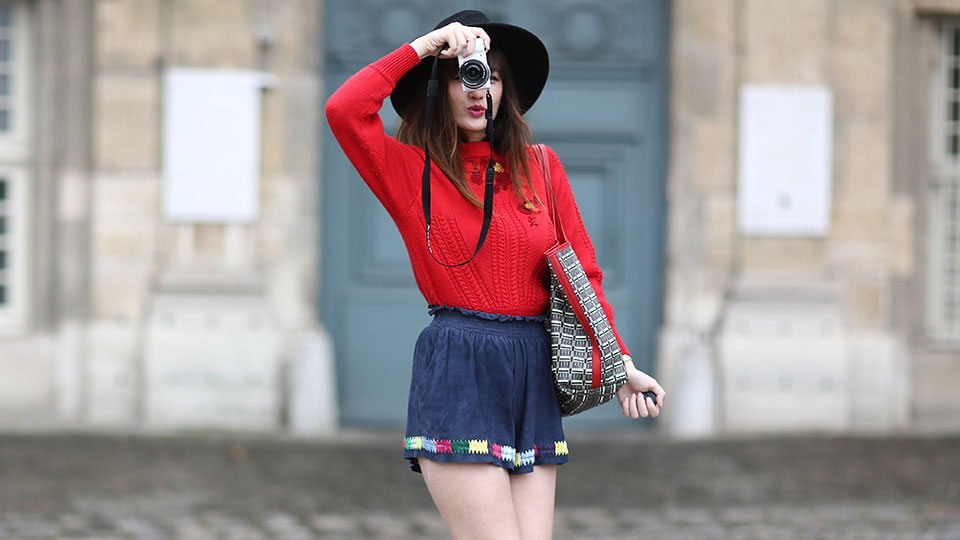 A woman in a red long sleeve shirt, blue shorts and black hat poses for the camera while holding a camera in her hand